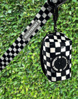 Dog poop bag holder in black and white checkered print with matching leash and carabiner clip against green hedge