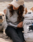 Woman sitting with two dogs wearing rescue gray hoodies, one with a star design and the other with a humorous message.