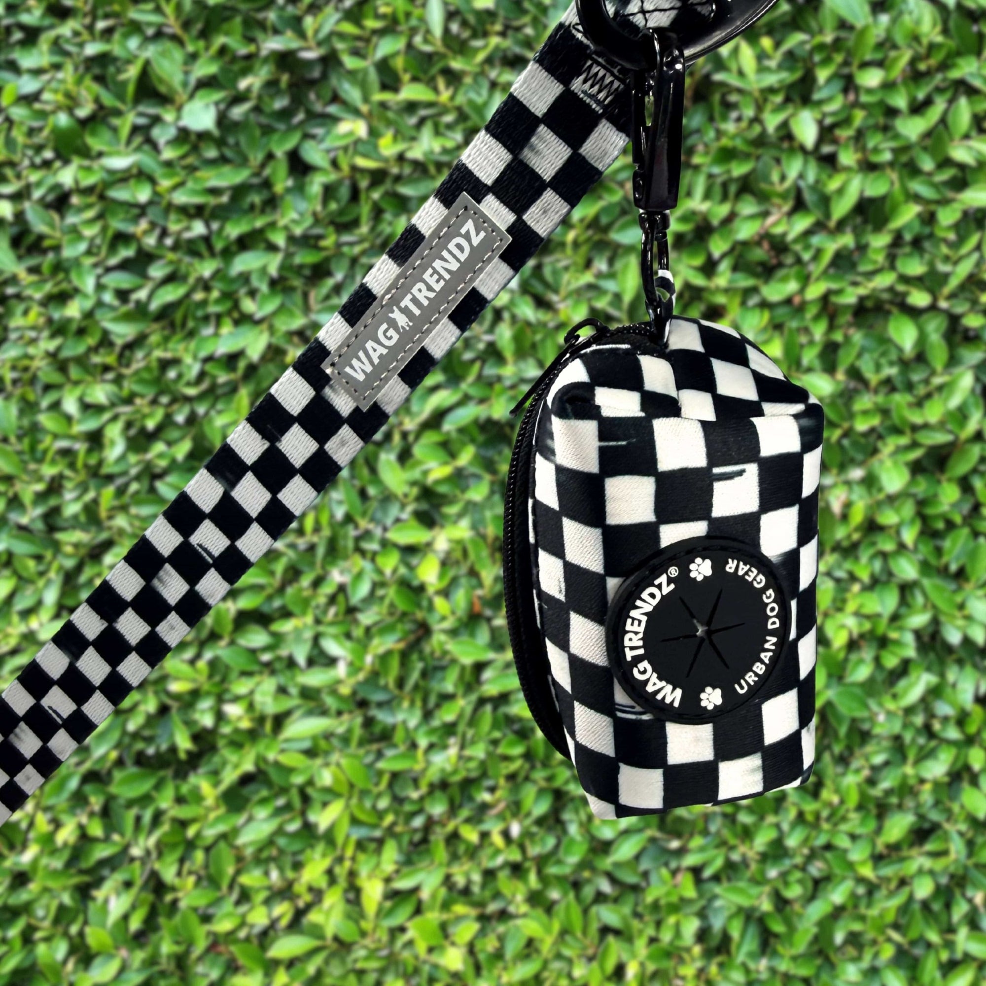 Dog poop bag holder in black and white checkered print with matching leash and carabiner clip against green hedge