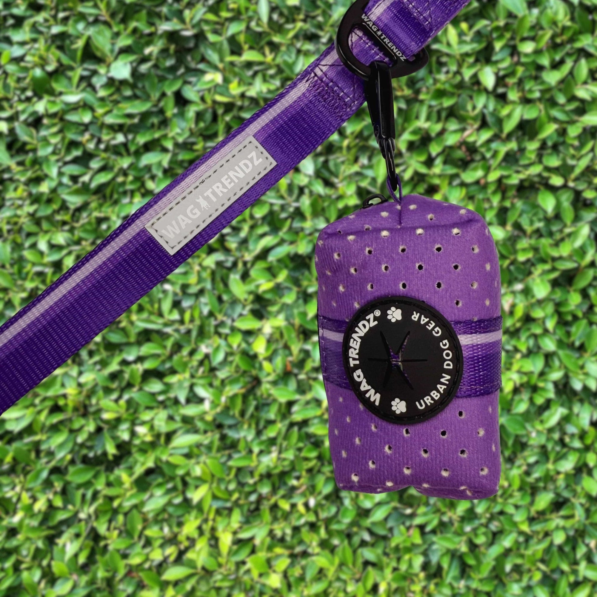 NeoCool™ Sport Poop Bag Holder in Purple with black logo dispenser attached to a leash against a green hedge background