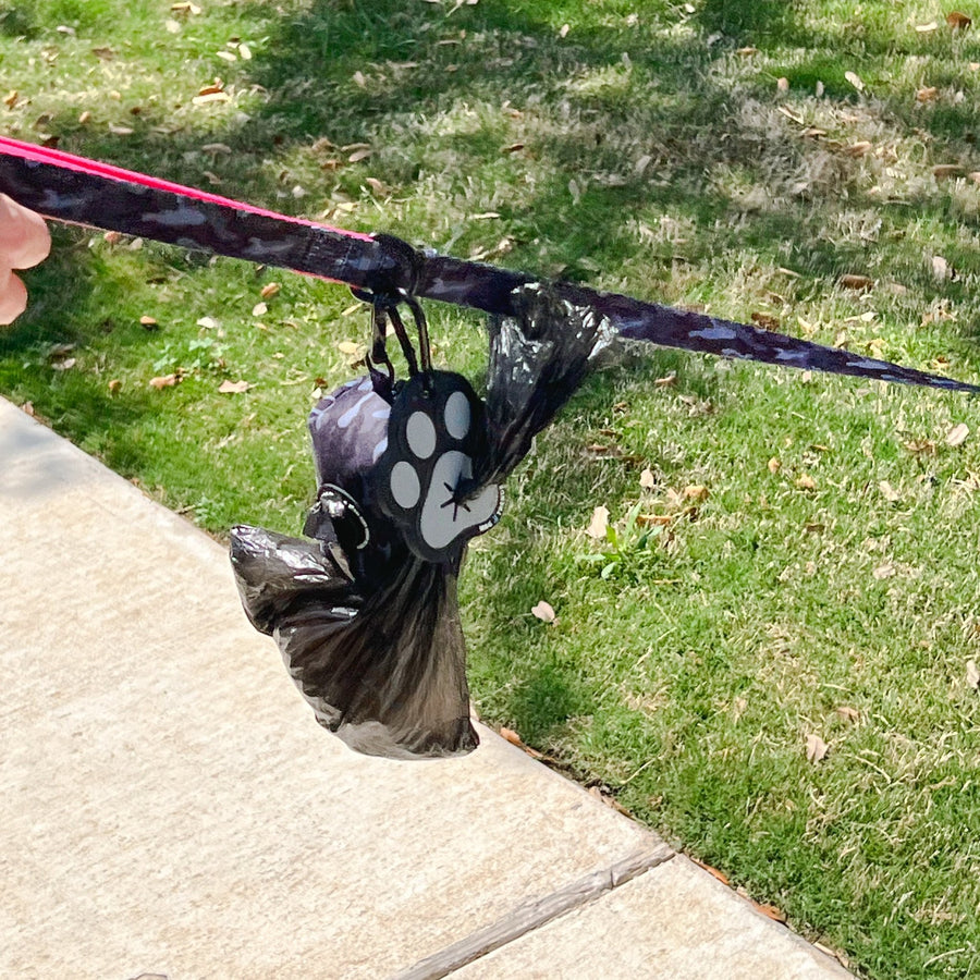 Poop Buddy - black and gray resin dog paw - hanging on a dog leash - outdoors with grass and sidewalk in background - Wag Trendz